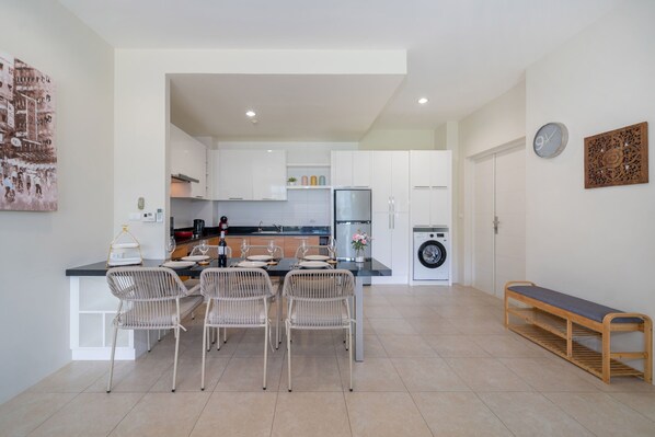 A modern kitchen with white cabinets, a dining area with wicker chairs, a built-in washing machine, and a bench.