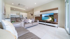 A modern living room with a large flat-screen TV, a beige sofa, and a kitchen in the background.
