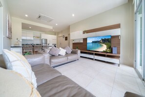A modern living room with a large flat-screen TV, a beige sofa, and a kitchen in the background.