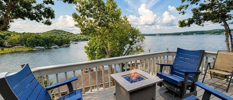 A deck with outdoor seating and a fire feature, overlooking a lake with boats and greenery.