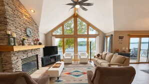 Un salón amplio con chimenea de piedra, televisor y ventana grande con vista a un cuerpo de agua.
