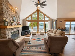 Un salón amplio con chimenea de piedra, televisor y ventana grande con vista a un cuerpo de agua.
