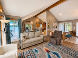 A spacious living area with a sofa, armchair, and a large rug, leading to a kitchen and dining area with wooden beams and a view of the outdoors.