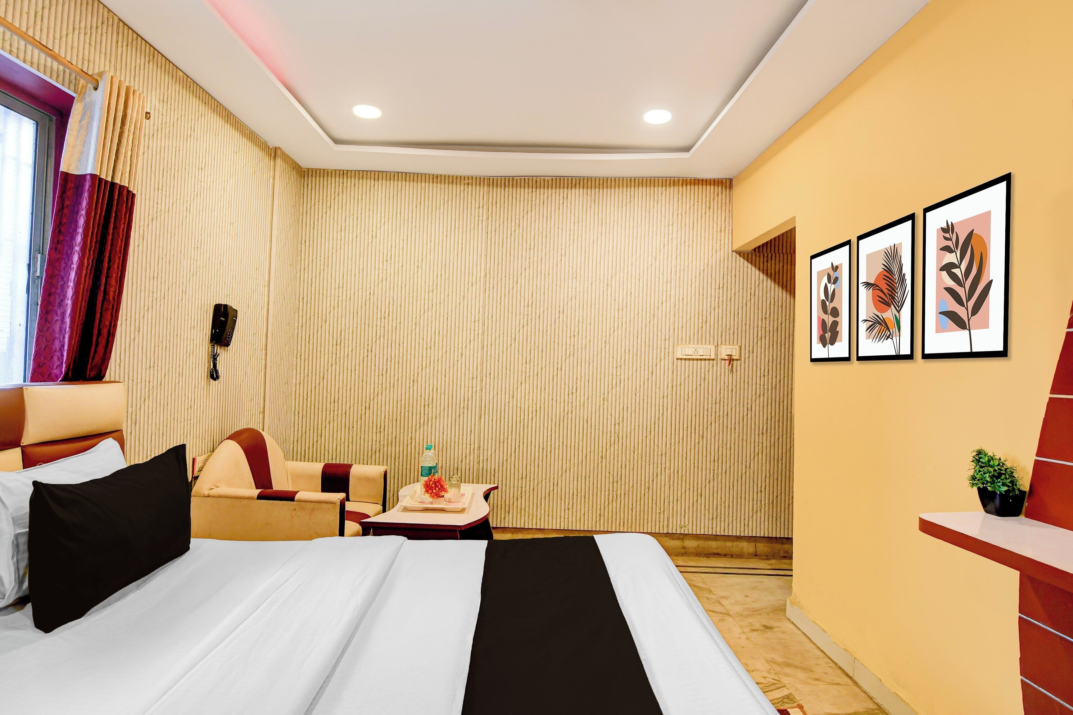 A hotel room with a bed, a desk, a chair, a small table with a bottle and a glass, a wall-mounted phone, and framed artwork on the wall.