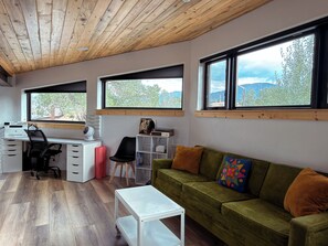 Een moderne kamer met een houten plafond, een groene bank met oranje kussens, een wit bureau met stoel, en grote ramen met uitzicht op bomen en bergen.