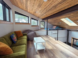 A modern loft bedroom with a sloped ceiling, large windows, a green sofa, and a small white table. - Lofted home in a quiet neighborhood of downtown Salida. California king bed plus super comfortable queen murphy bed and FullXL floor mattress in loft. Two levels, two bathrooms, beautifully landscaped private courtyard with hot tub and gas fire pit. (Salida)