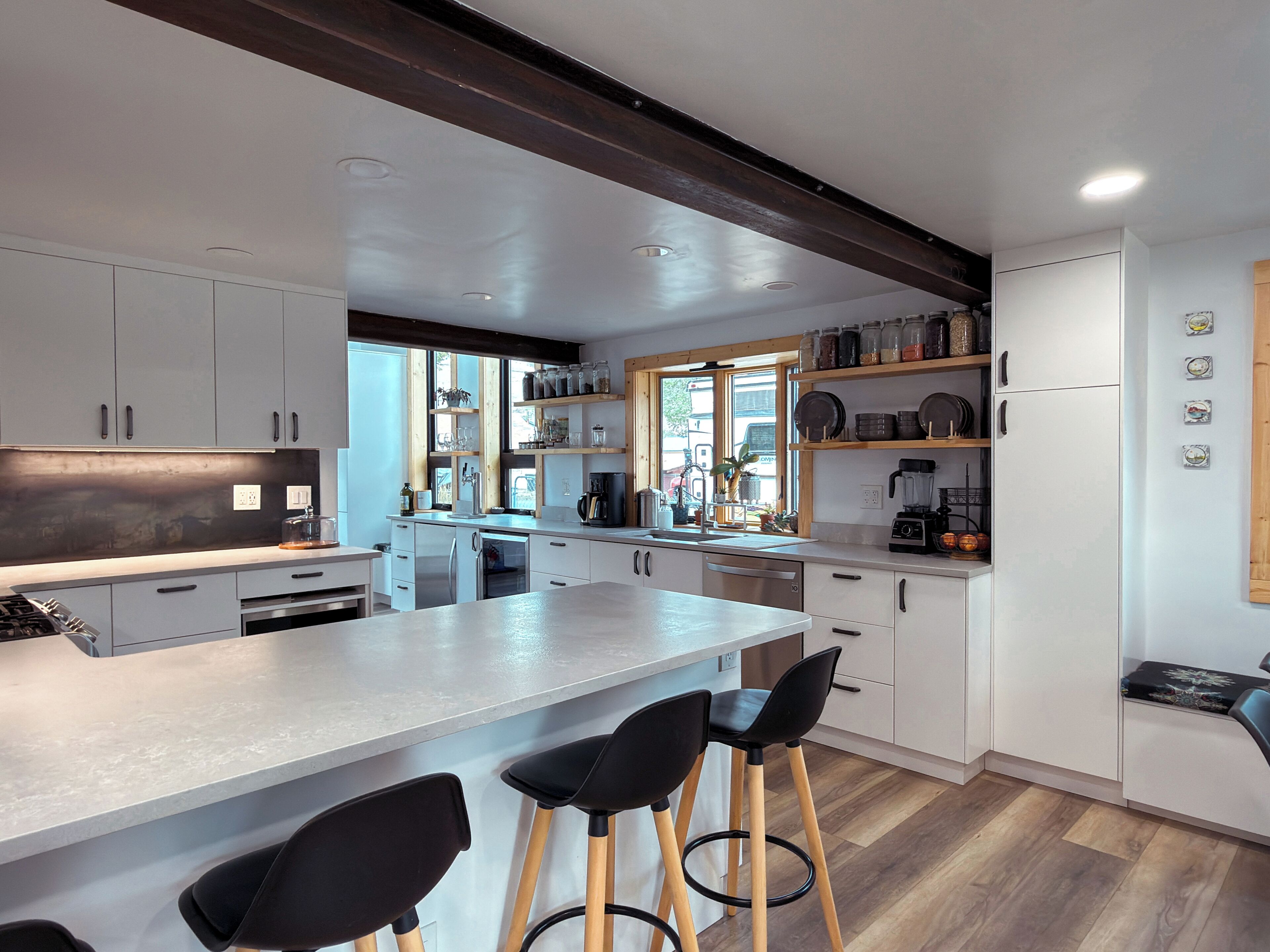 Une cuisine moderne avec des meubles blancs, un îlot central doté de tabourets de bar et une poutre en bois au plafond.