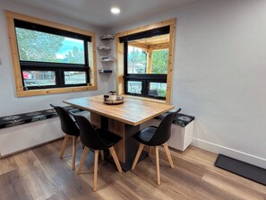 Ein Zimmer mit einem Holztisch, vier Stühlen und zwei Fenstern mit Holzrahmen.