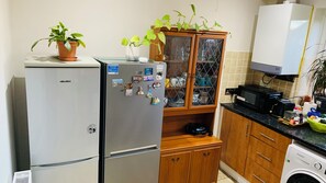 A compact kitchen with a refrigerator, a microwave, a washing machine, wooden cabinets, and a glass-fronted cabinet with plants. - A Clean & Cosy Abode for a Comfortable Homestay (Sutton)