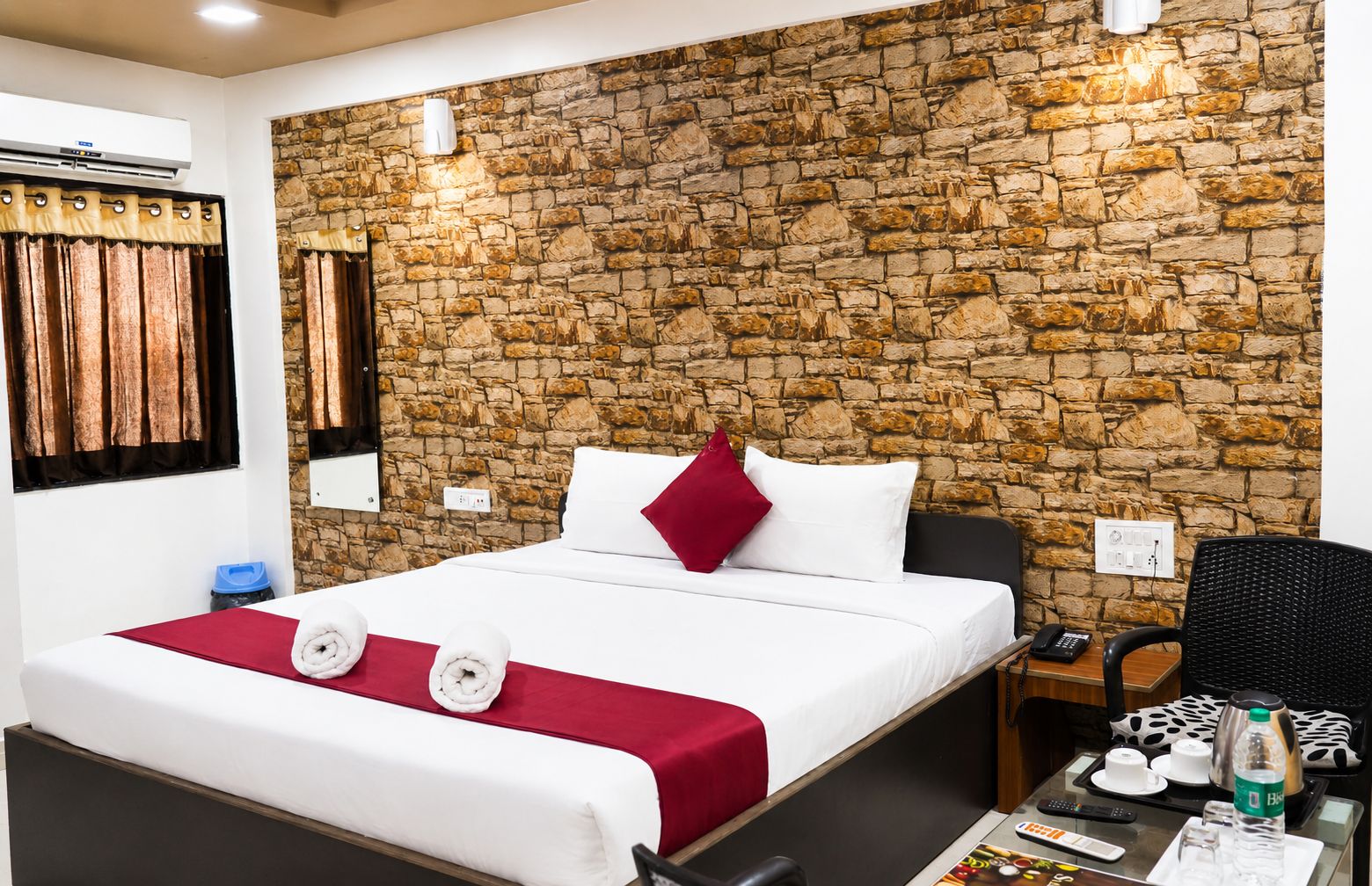 A hotel room with a stone wall, a bed with white bedding and a red pillow, a bedside table with a phone and a bottle, and a chair.