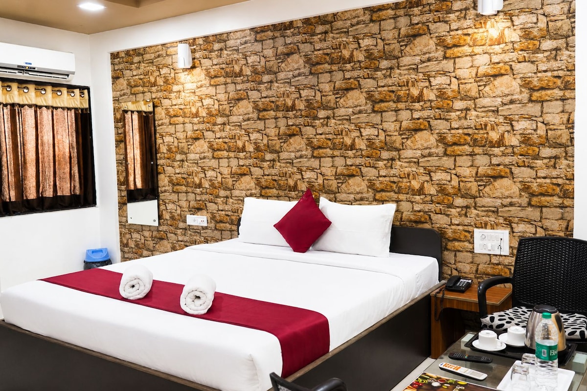 A hotel room with a stone wall, a bed with white bedding and a red pillow, a bedside table with a phone and a bottle, and a chair.