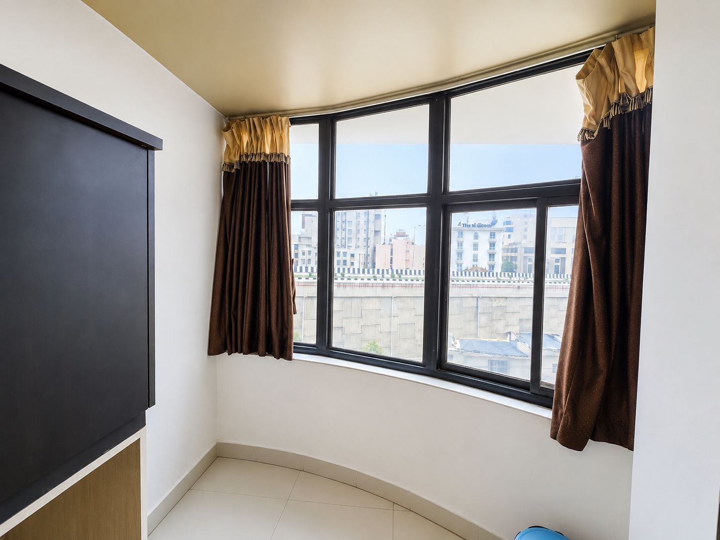 Una habitación con un mueble negro grande, un ventanal curvo con cortinas y vistas a edificios.