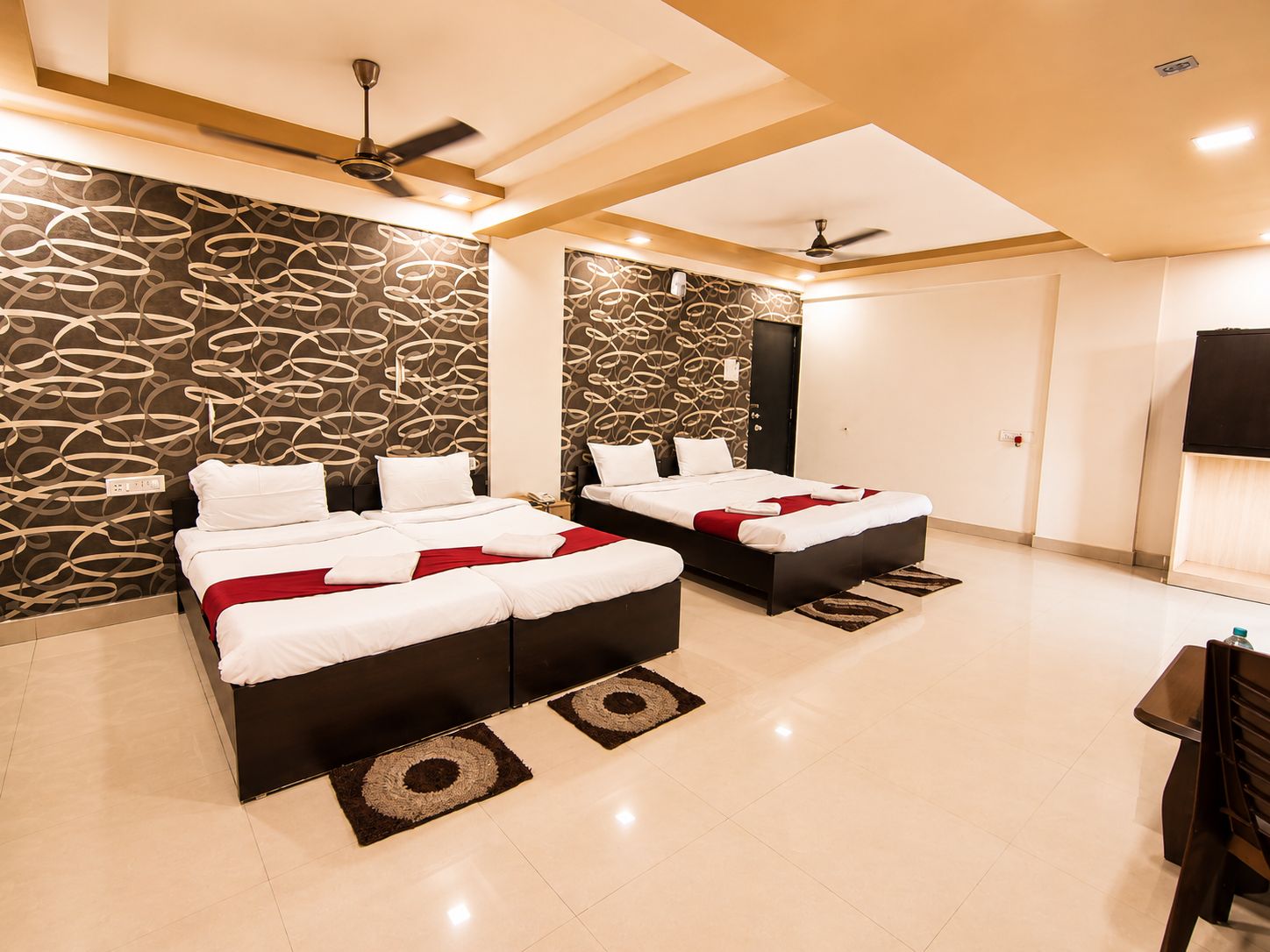 A hotel room with two beds, a flat-screen TV, ceiling fans, and a patterned wall.