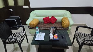 A room with a green sofa, two patterned chairs, a glass coffee table with a tray of items, and a black laptop on a stand. - Hotel Sukh Sagar (Veraval)