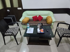 Una habitación con un sofá verde, dos sillones estampados, una mesa de centro de vidrio con una bandeja de objetos y un portátil negro sobre un atril.