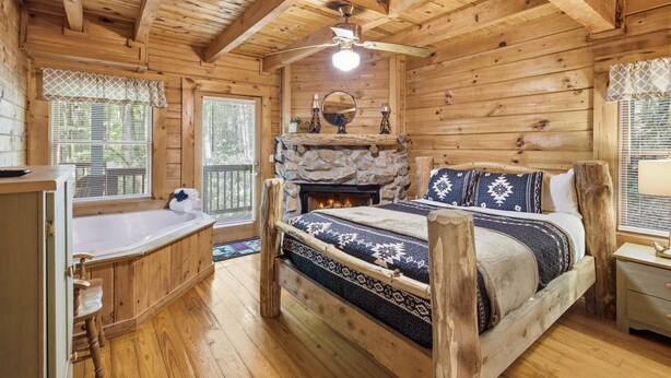 A log cabin interior with a stone fireplace, a bed with patterned bedding, a wooden bathtub, and a ceiling fan. - 3 Brothers Bears | Smoky Mountain Escape (Sevierville)