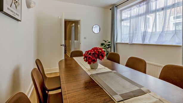 Ein Essbereich mit einem langen Holztisch, Stühlen, einer Vase mit roten Blumen und einem Fenster mit durchsichtigen Vorhängen.