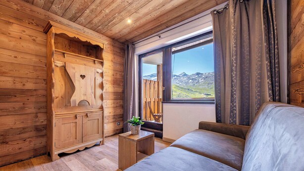 Ein Holzinnenraum mit einer Couch, einem Holzkabinett mit Herzförmigem Ausschnitt und einem Fenster mit Blick auf die Berge.