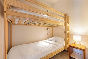 A bunk bed with a wooden frame, a single bed below, a nightstand with a lamp, and a wall-mounted light fixture. - “V5” by Versant Collection (Val Thorens)