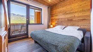 Ein Schlafzimmer mit Holzbalkendecke, einem Bett mit weißer Bettwäsche, Blick auf Berge durch eine Schiebetür und einem kleinen Tisch mit Stuhl.