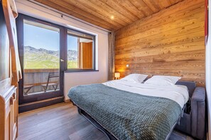 A bedroom with a wooden ceiling, a bed with white bedding, a view of mountains through a sliding door, and a small table with a chair. - Residence Les Olympiades, Ski In-Out with WiFi in Val Thorens, “V5” by Versant Collection (Val Thorens)