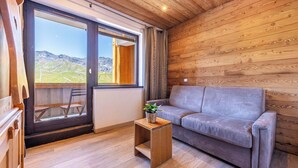 Ein Holzinnenraum mit einer Couch und einem kleinen Tisch, eine Schiebetür zu einem Balkon und ein Blick auf eine gebirgige Landschaft.