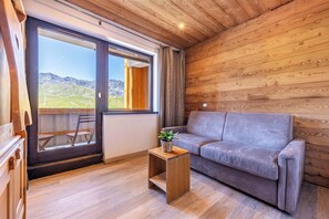 A wooden interior with a sofa and a small table, a sliding glass door leading to a balcony, and a view of a mountainous landscape. - Residence Les Olympiades, Ski In-Out with WiFi in Val Thorens, “V5” by Versant Collection (Val Thorens)