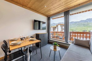 A room with a wooden ceiling, a dining table set with plates and glasses, a TV mounted on the wall, and a sofa with a view of a mountainous landscape. - “V13” by Versant Collection (Val Thorens)