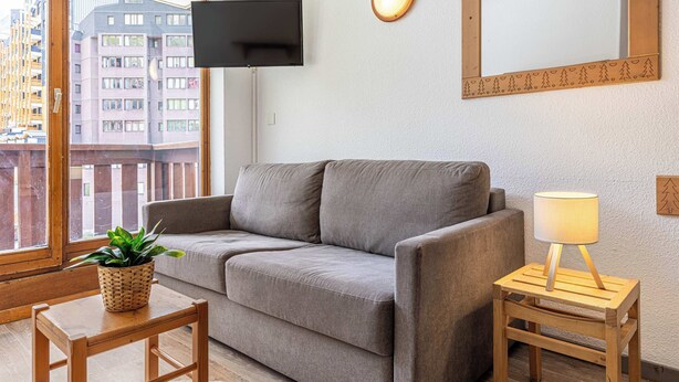 Ein modernes Wohnzimmer mit einem grauen Sofa, einem kleinen Holztisch mit Pflanze, einer Stehlampe und einem an der Wand montierten Fernseher.