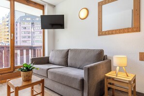 A modern living room with a grey sofa, a small wooden table with a plant, a floor lamp, and a TV mounted on the wall. - "V10” by Versant Collection (Val Thorens)