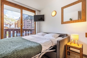 A hotel room with a bed, a bedside table with a lamp, a flat-screen TV mounted on the wall, and a large window offering a view of buildings and a mountain. - "V10” by Versant Collection (Val Thorens)