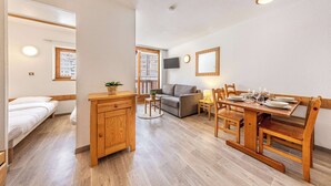 Ein kompakter Wohnraum mit Bett, Holzschrank, Sofa, Esstisch mit Stühlen und einem an der Wand befestigten Fernseher.