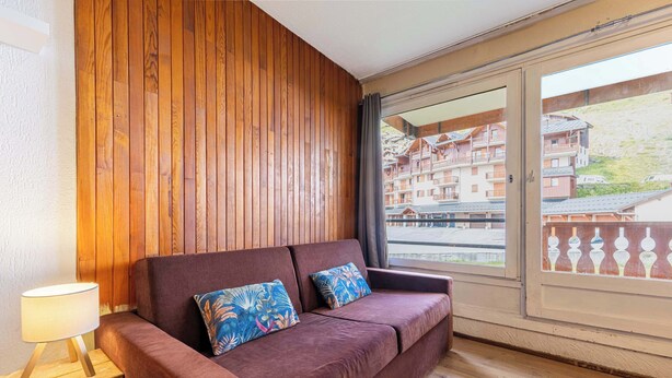 A room with a wooden panel wall, a brown sofa with floral cushions, a small table with a lamp, and a sliding glass door leading to an outdoor area with buildings.