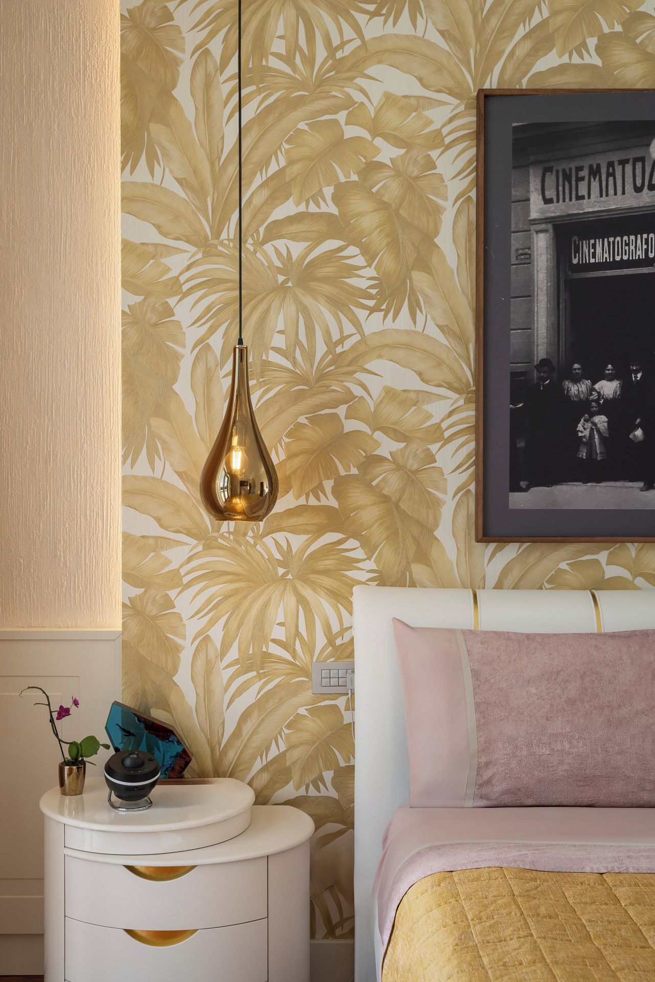A bedroom with a bed, a nightstand, a wall-mounted light, a framed picture, and a tropical wallpaper.