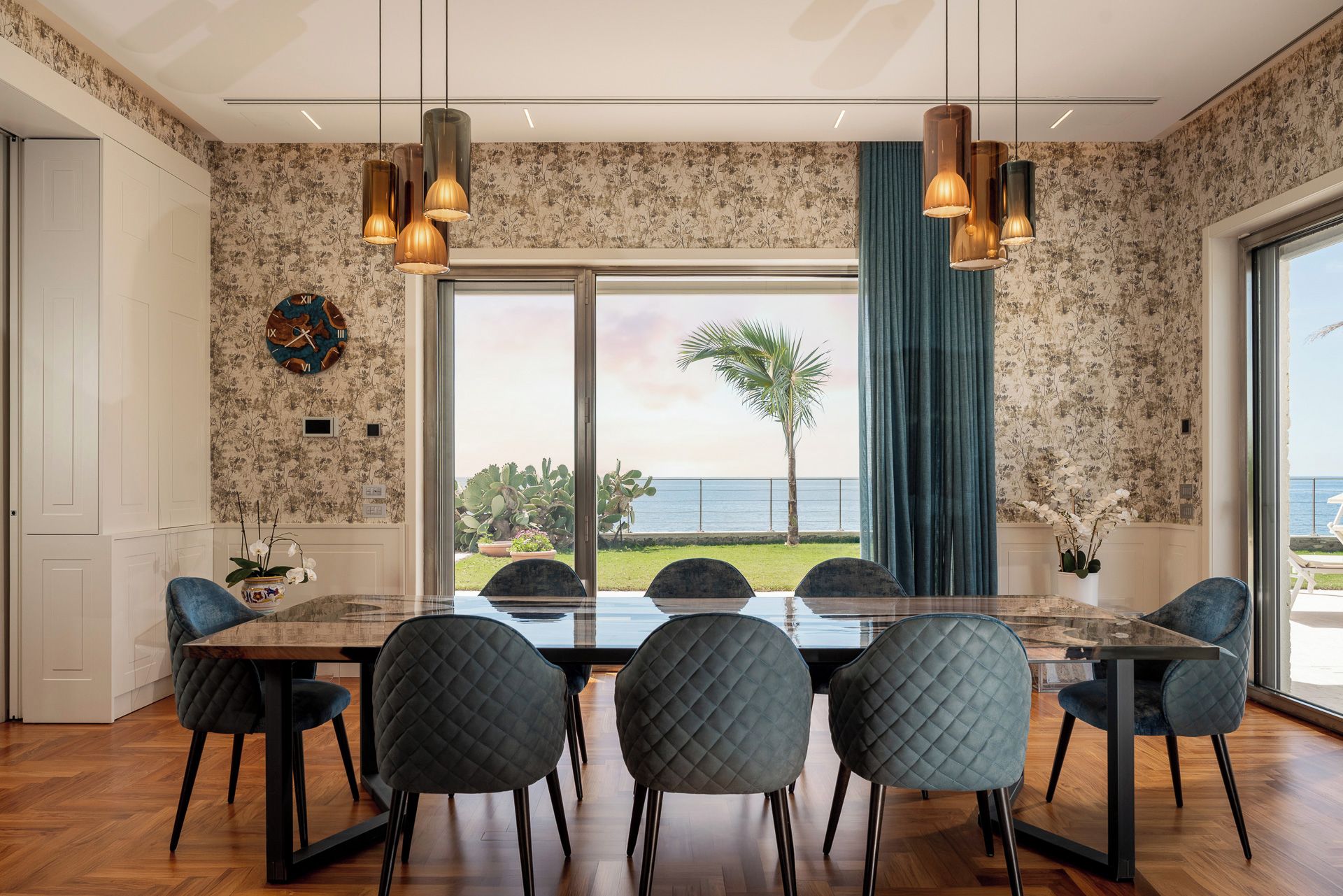 A dining area with a large table and six chairs, a view of the ocean, and a tropical plant outside.