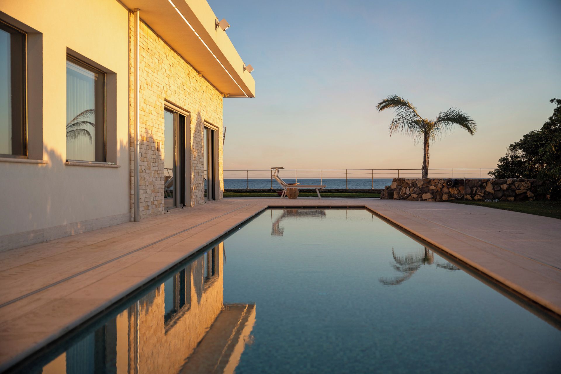 Une piscine offrant une vue dégagée sur la mer, un mur en pierre et un palmier.