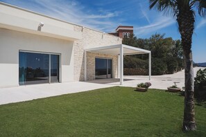 A modern house with a stone facade, large sliding glass doors, a covered patio area, and a well-maintained lawn.