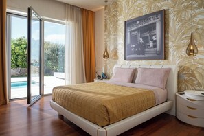 Ein Schlafzimmer mit Bett, Nachttisch, Lampe, einem gerahmten Bild und Blick auf einen Pool.