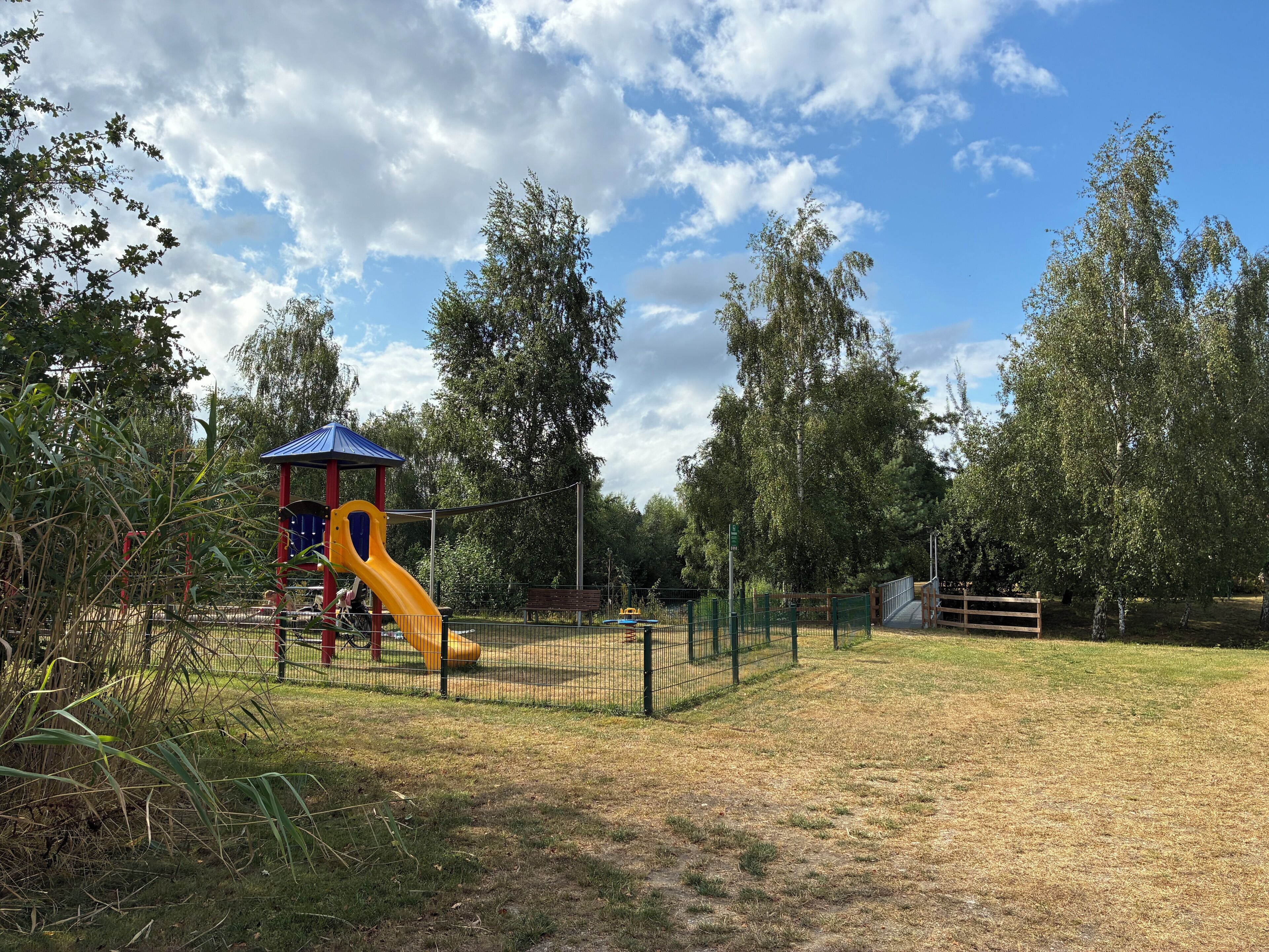 Un parque infantil con un tobogán amarillo y una estructura de juegos roja y azul, rodeado de árboles y una zona vallada.