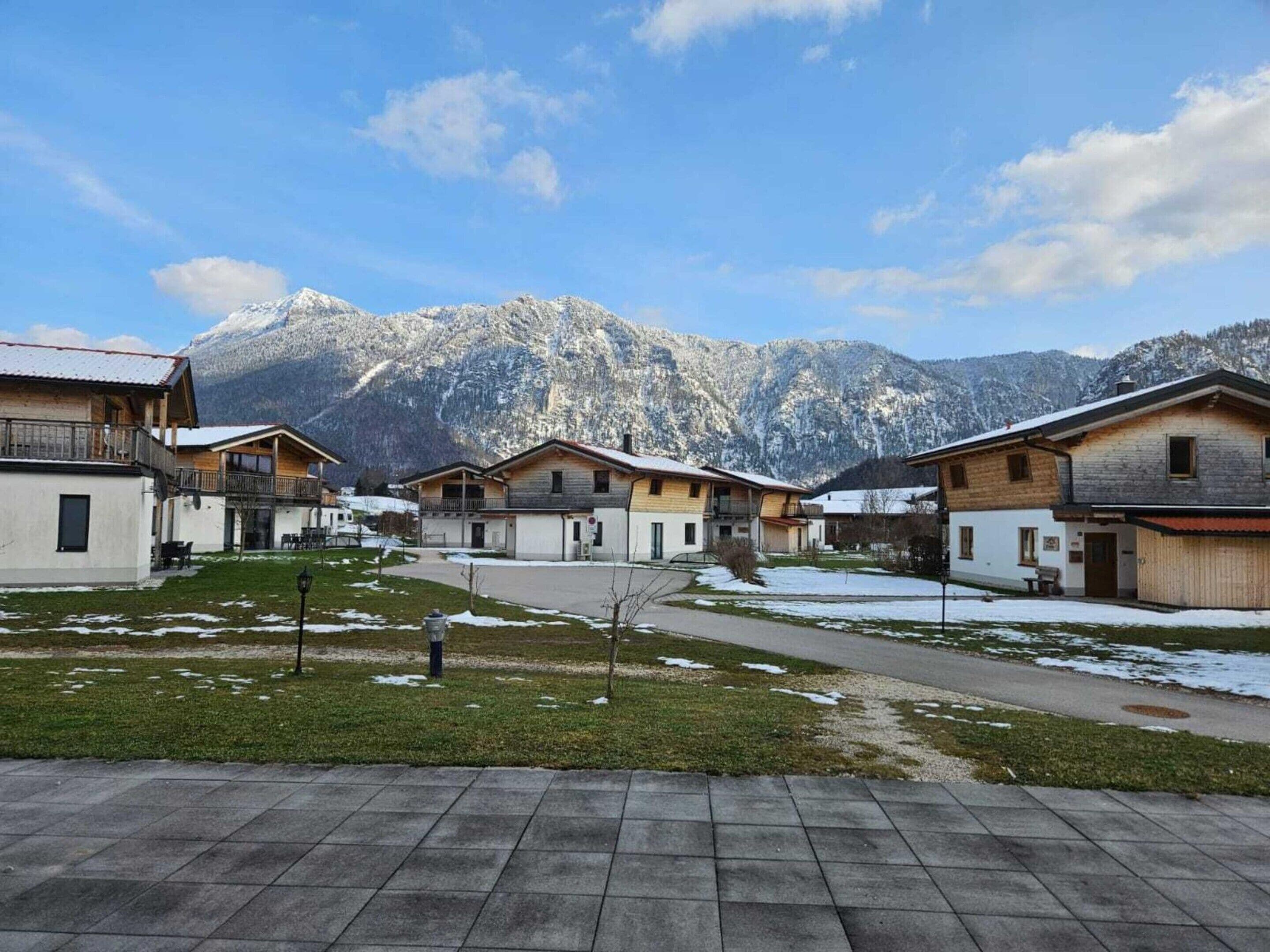 Reihenweise Holzhäuser vor einer Bergkulisse, ein gepflasterter Weg und eine kleine, grasbewachsene Fläche mit vereinzelten Schneeflecken.