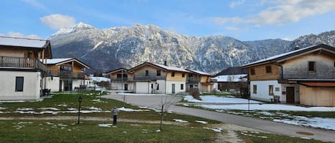 Une rangée de bâtiments en bois, avec une chaîne de montagnes en arrière-plan, un chemin pavé et une petite zone herbeuse parsemée de neige.