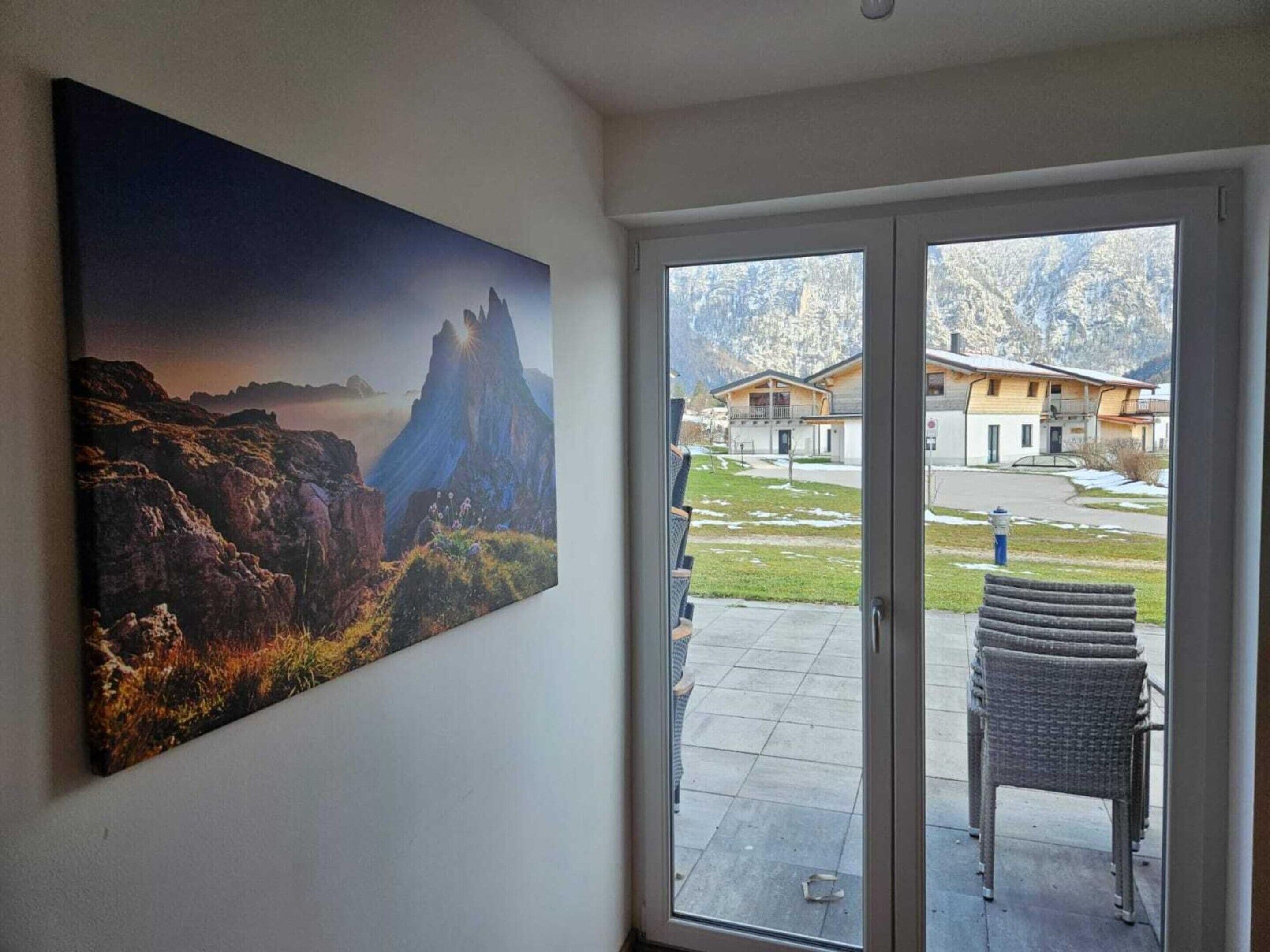 Ein Zimmer mit großem Fenster, einem Berglandschaftsbild und Blick auf eine Wohngegend mit Häusern und blauem Himmel.