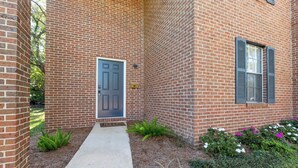 Un bâtiment en briques avec une porte centrale, un petit chemin menant à cette porte et une fenêtre avec des volets.