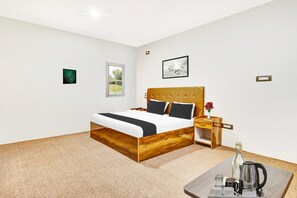 Habitación de hotel con una cama de madera, una mesita de noche con lámpara, una pequeña mesa con hervidor y una ventana con vista a zonas verdes.