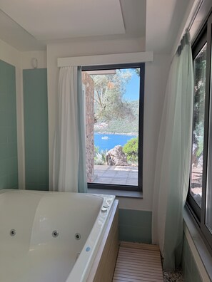 A bathroom with a bathtub, a window with curtains, and a view of a rocky landscape and water body.