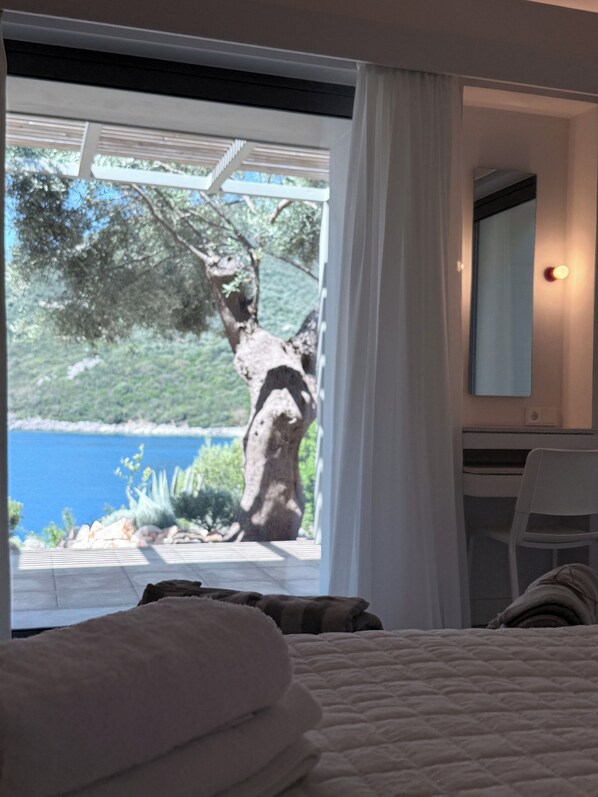 A bedroom with a bed, a chair, a desk, a window with curtains, and a view of a tree and water outside.