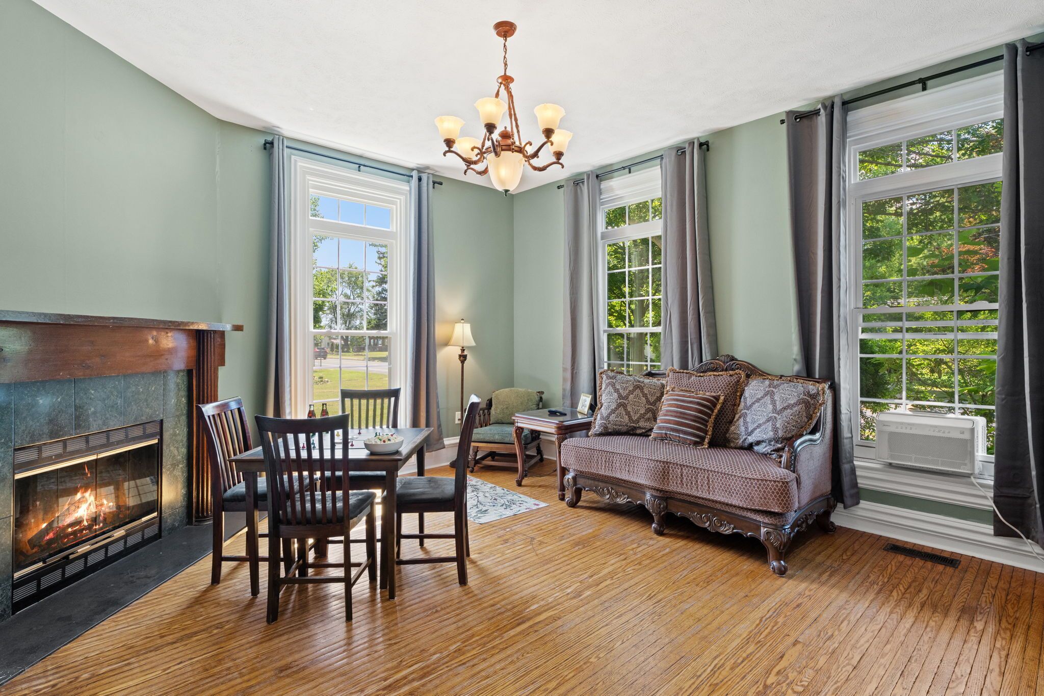 A living room with a fireplace, a sofa, a dining table with chairs, and large windows with curtains.