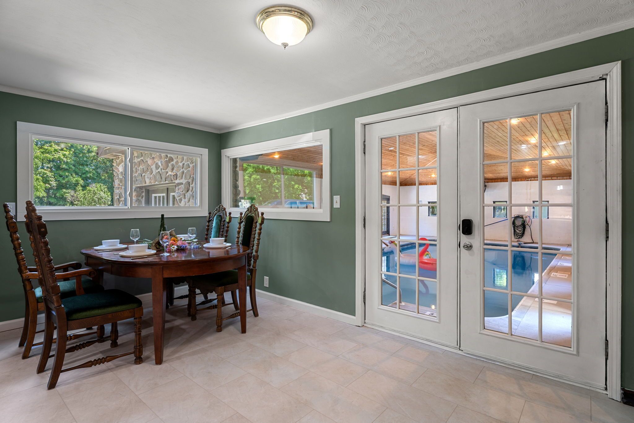 A dining area with a table and chairs, a view of a swimming pool through a glass door, and a stone wall with a window.