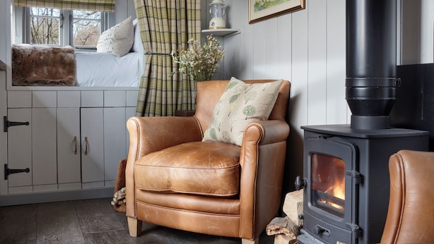 A cozy living room with a leather armchair, a wooden cabinet, a fireplace, and a window with curtains. - Hilltop Shepherds Hut with Hot Tub and Tennis (Hambleden)