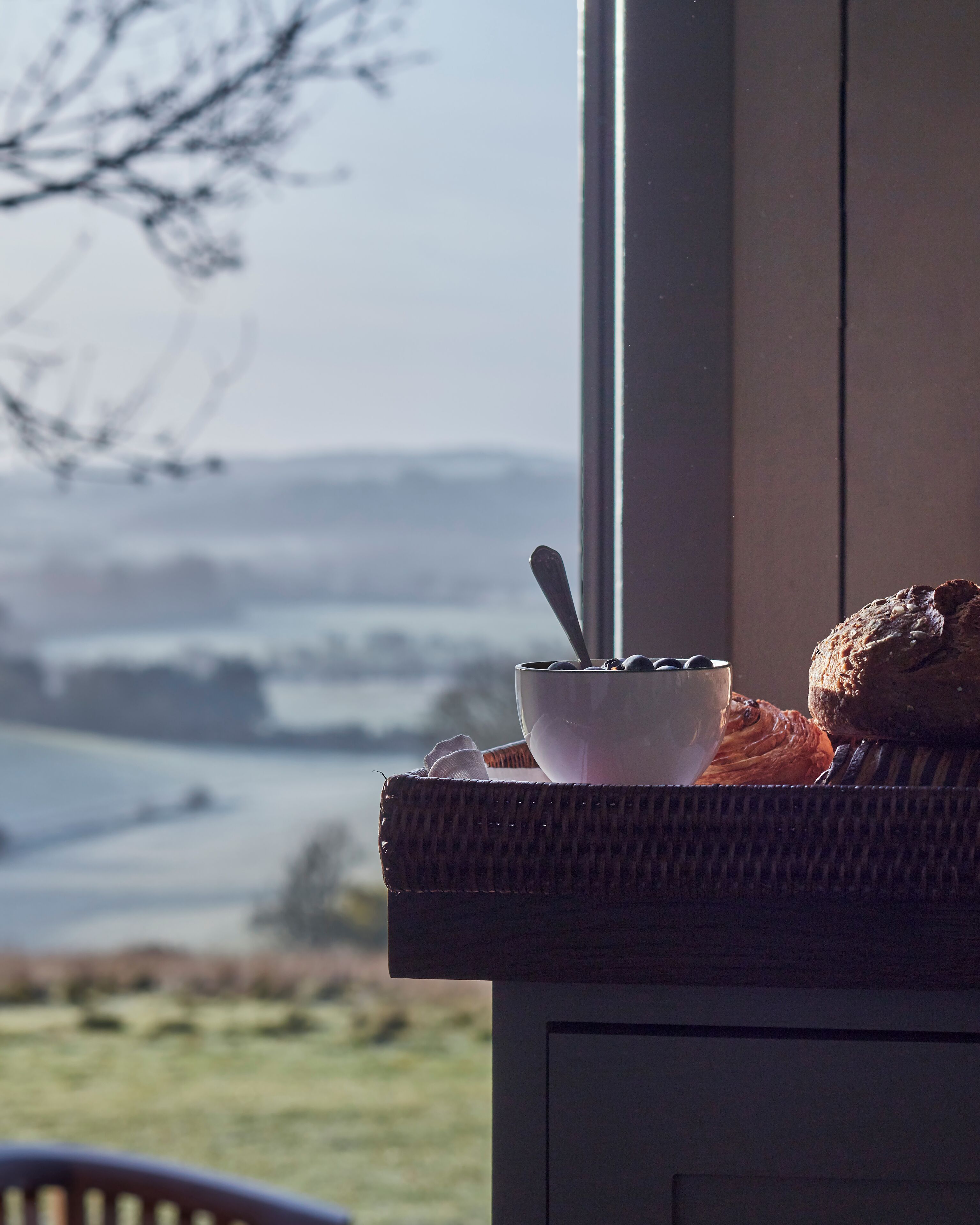 Un desayuno con un tazón de cereal y una hogaza de pan sobre una mesa cerca de una ventana que da a un paisaje.